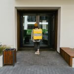 A delivery person with a yellow thermal bag approaches a customer's door in a city setting.